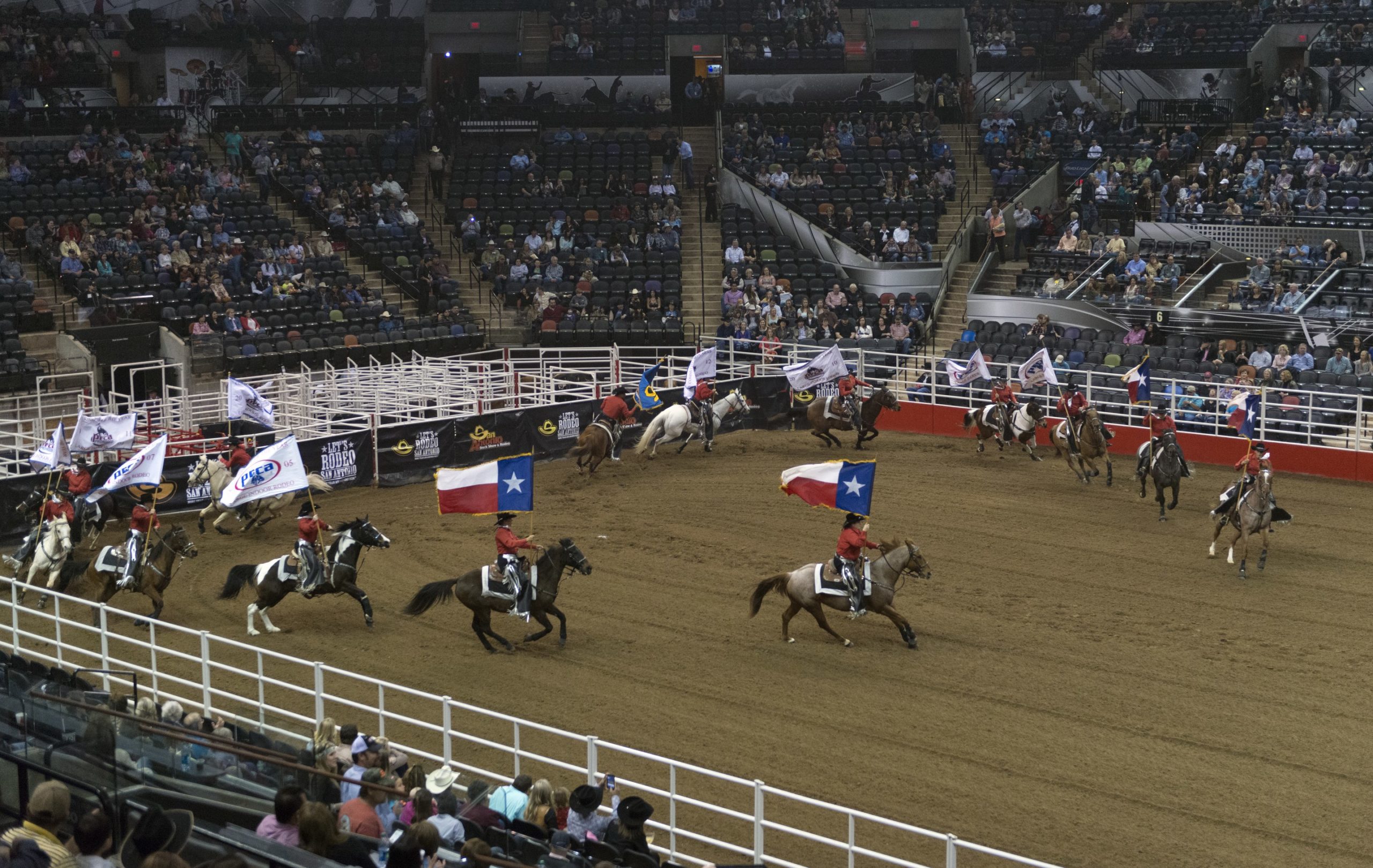 Here is a picture of some Bronc Riders at the Rodeo, waving Texan flags. A large crowd is watching and cheering,